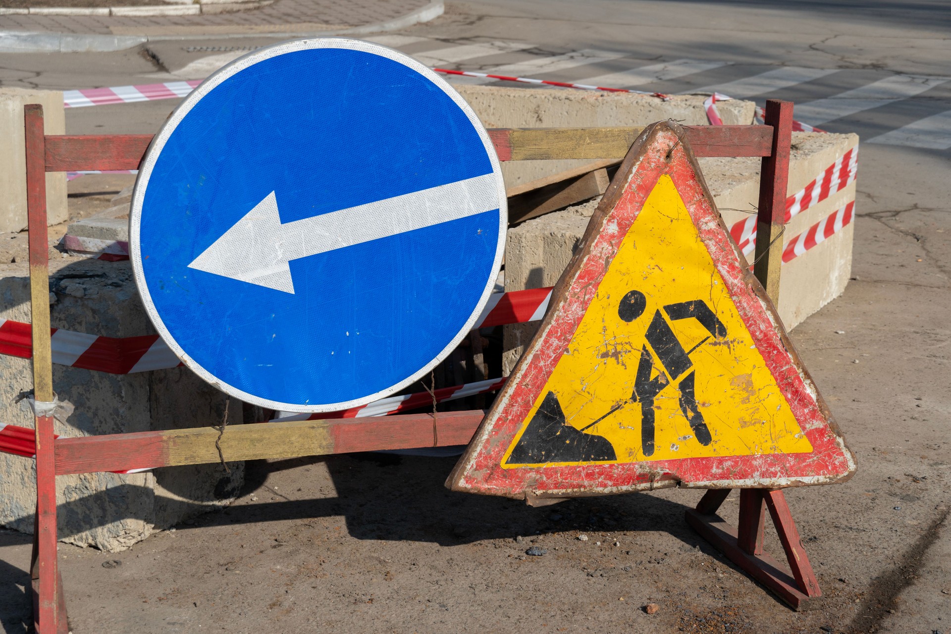 Two traffic signs stand along a city street, one directing left and the other warning of construction work. Bright sunlight illuminates the scene